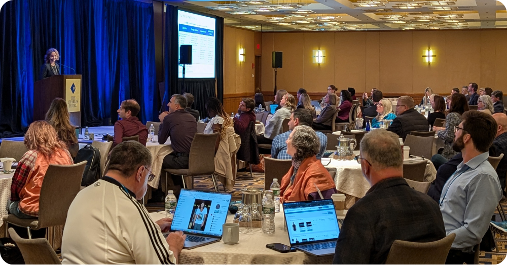 A speaker presents at a podium to an audience seated at round tables in a conference room at The Charles Hotel.