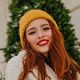 A smiling young woman with red hair, wearing a yellow knit beanie and a white coat, outdoors.